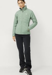 Chaqueta acolchada verde claro con costuras verticales, cuello alto y cierre de cremallera. Pantalones negros ajustados con bolsillos con cremallera y costuras reforzadas.