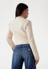 Beige umělá kůže bunda s vysokým límcem, úzkým střihem a viditelnými švy. Nosená s modrými džíny z denimu s trojúhelníkovým logem.