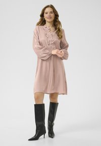 Vestido rosa claro con bordados florales en el escote, mangas fluidas y un corte relajado, combinado con botas negras de punta alta hasta la rodilla.