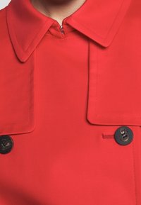 Manteau rouge avec un col pointu, doté d'une fermeture éclair cachée et de deux boutons foncés, texture lisse et design structuré.