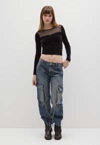 Zwart shirt met lange mouwen en een doorschijnend inzetstuk bij de halslijn, gecombineerd met losse, vervaagde blauwe cargobroeken met grote zakken en ritssluitingdetails.