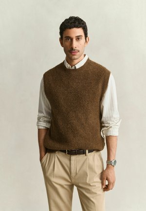 Man met kort krullend haar die een bruine mouwloze trui draagt over een wit gestreept overhemd en beige broek, staand met één hand in de zak.