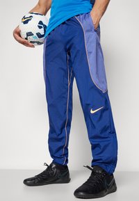 Blaue Nike Sporthosen mit glänzender Textur, lila seitlichen Akzenten und kontrastierenden orangen Biesen. Schwarze sportliche Schuhe und ein Fußball sind vorhanden.