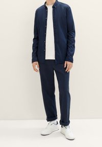 Camicia blu navy a maniche lunghe sopra una t-shirt bianca, abbinata a pantaloni blu navy e sneaker bianche. Tessuto di cotone, vestibilità ampia, maniche arrotolate.