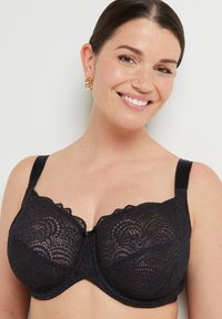 Reggiseno in pizzo nero con motivi floreali, coppe con ferretto, spalline regolabili e bordi scallopati; chiusura centrale con gancio per supporto.