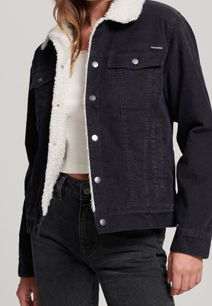 Frau trägt schwarze, geknöpfte Jeansjacke mit weißem Sherpa-Futter über einem weißen Cropped-Top und schwarzen Jeans.