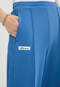 Blauwe trainingsbroek van gladde stof heeft een enkele zijzak en een geborduurd logo op de dij. Gedetailleerd met plooien.