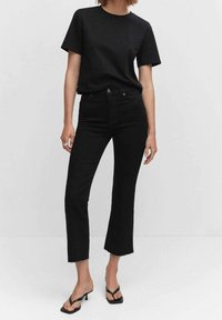 Nainen, joka käyttää yksinkertaista mustaa t-paitaa, korkeavyötäröisiä mustia farkkuhousuja, joissa on raaupistetut helmat, ja mustia korkokenkiä, seisoo valkoista taustaa vasten.