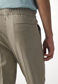 Gros plan d'une personne portant un pantalon beige avec une taille élastique et une poche passepoilée à l'arrière, main posée sur le côté.