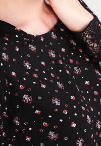 Tessuto nero con un motivo floreale in rosso e bianco. Collo tondo e dettagli in pizzo sulle maniche, con piccole forme distribuite uniformemente.