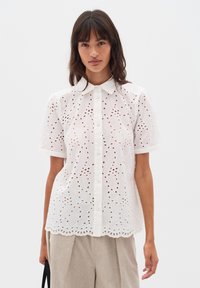 Blouse bianca a maniche corte con occhielli ricamati e orlo ondulato, caratterizzata da una chiusura con bottoni e un colletto classico. Tessuto morbido e leggero.