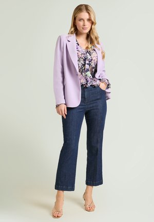 Young woman stands with hand in pocket wearing a lilac blazer, floral blouse, dark blue jeans, and gold open-toe heels on a plain background.