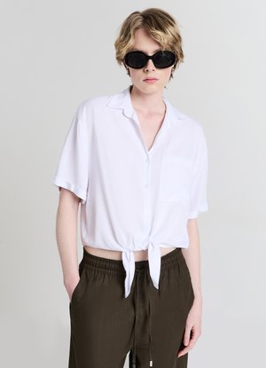 Blouse blanche à manches courtes avec nouage au devant et une poche ; associée à un pantalon vert foncé à taille élastique. Tissu lisse et léger.