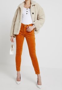 Beżowa kurtka z sztucznego futra, biała bluzka z prążkami z guzikami, pomarańczowe spodnie z korduroju i białe szpilki, trzymająca małą białą torebkę.