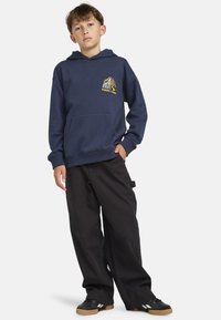 Hoodie marine avec une poche kangourou et des détails graphiques jaunes sur la poitrine, associé à un pantalon large noir et des baskets noires.