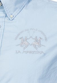 Ljusblå skjorta med knappar som har en broderad poloscen med två ryttare och hästar, samt texten "LA MARTINA" nedtill.