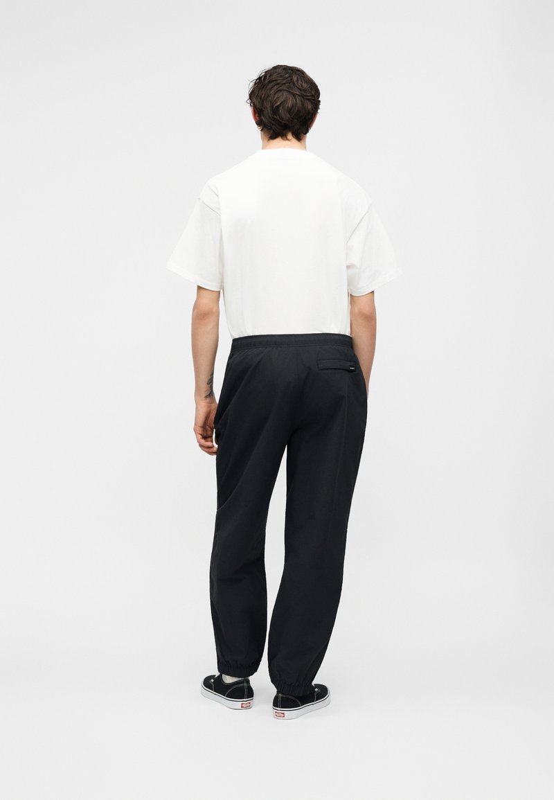 Mann von hinten gesehen, trägt ein weißes kurzärmeliges Hemd, schwarze Hosen mit elastischem Bund und schwarze Sneakers mit niedrigem Schaft vor weißem Hintergrund.
