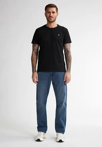 Černé tričko s kulatým výstřihem, doplněné světle modrými džínami. Pletená látka, volný střih a viditelné detaily tetování na pažích. Bílé tenisky.