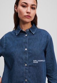 Tamsiai mėlyna džinsinė marškinė su sagų priekio ir apykakle. Yra sidabriniai aksesuarai ir siuvinėtas tekstas "KARL LAGERFELD JEANS."