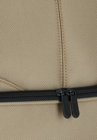 Bolsa de tela beige con una superficie texturada, con cremalleras y costuras negras en contraste. Los tiradores de la cremallera están etiquetados como "DERMATA."