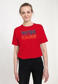 Ung kvinde med lys hud og blondt hår iført en rød WSQK The Squawk 94.5 FM t-shirt og sorte bukser, stående mod en hvid baggrund.
