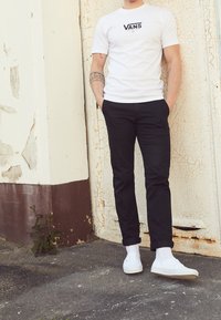 Osoba w białej koszulce Vans, czarnych podwiniętych spodniach i białych sneakersach stoi z rękami w kieszeniach na tle popękanej, łuszczącej się ściany.
