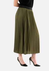 Jupe plissée vert olive avec une ceinture texturée noire, au design fluide et longueur mi-mollet, associée à des escarpins noirs à talons pointus.