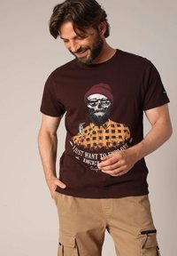 Camiseta corta marrón de manga corta con un estampado gráfico de una calavera que lleva gafas de sol y un gorro, con un diseño de camisa de cuadros.