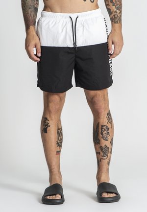 Uomo che indossa pantaloncini da bagno bianchi e neri con testo laterale, sandali neri a ciabatta e tatuaggi visibili su braccia e gambe, in piedi su uno sfondo semplice.