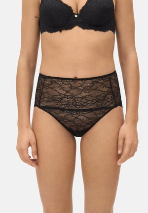 Ropa interior de encaje negro de talle alto y sujetador de encaje a juego, llevados por una persona con los brazos relajados a los lados, fondo neutro.