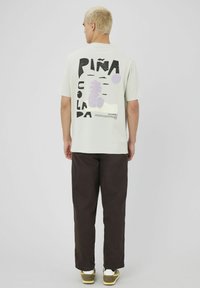 T-shirt vert clair avec un design graphique noir et violet et le texte "Piña Colada" au dos ; associé à un pantalon marron foncé.
