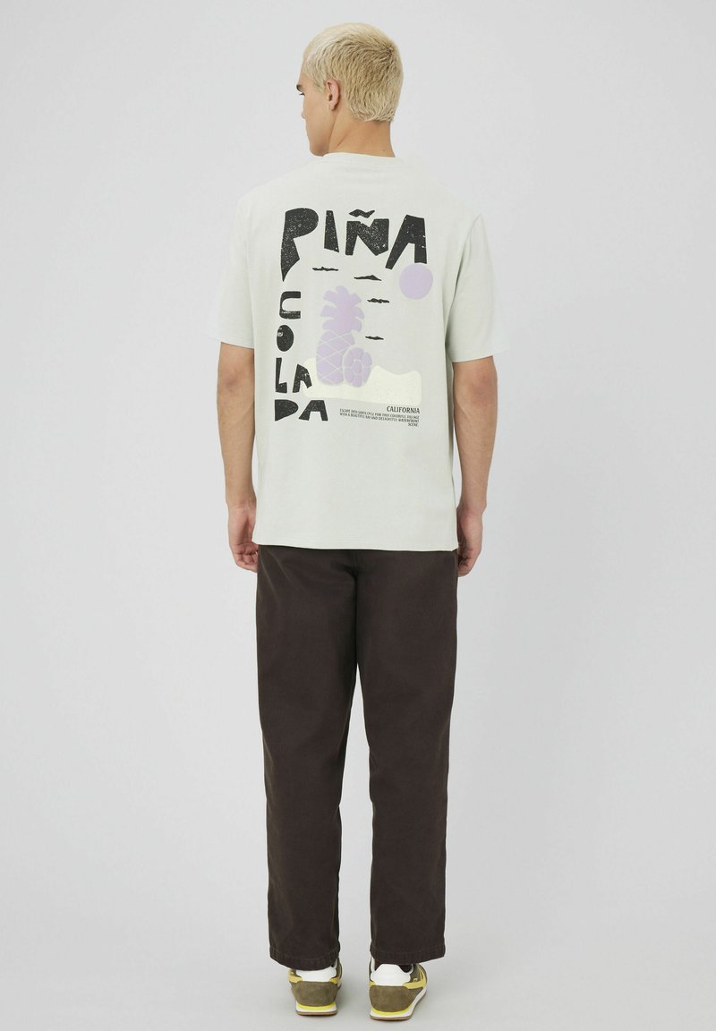 T-shirt vert clair avec un design graphique noir et violet et le texte "Piña Colada" au dos ; associé à un pantalon marron foncé.