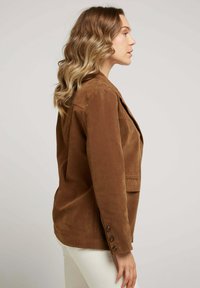 Blazer en daim marron avec une poche unique, des détails de poignet boutonné et une coupe ajustée, présenté de profil sur un fond uni.