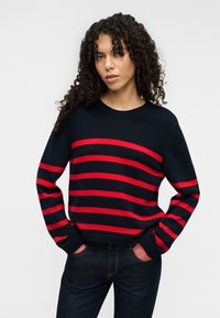 Πλεκτό πουλόβερ σε ναυτικό μπλε με οριζόντιες κόκκινες ρίγες, στρογγυλή λαιμόκοψη και ριμπ μανσέτες. Συνδυασμένο με σκούρο τζιν.