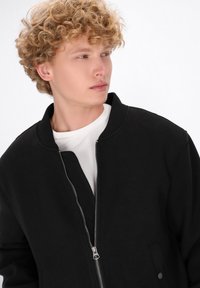 Giovane uomo con capelli ricci biondi, che indossa una giacca nera parzialmente zipata sopra una camicia bianca, mentre guarda di lato su uno sfondo bianco.