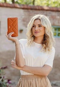 Hnědá peněženka z umělé kůže s prošívaným vzorem a logem. Žena ji drží proti rozmazanému venkovnímu pozadí, má na sobě bílé tričko a béžovou sukni.