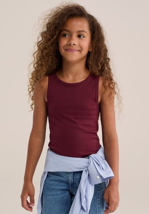 Junges Mädchen mit lockigem Haar, trägt ein ärmelloses burgunderfarbenes Top und blaue Jeans, mit einem hellblau gestreiften Hemd um die Taille gebunden.
