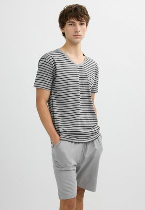 Joven con camiseta de rayas gris y negra con cuello en V y pantalones cortos gris claro, de pie con las manos en los bolsillos contra un fondo liso.