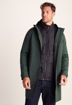 Man wearing dark green long coat over black quilted zip-up jacket and dark jeans, looking slightly to his right against plain background.