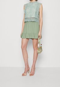 Abito a strati verde chiaro con dettagli intricati in pizzo sulla parte superiore, gonna svasata e orlo arruffato; abbinato a sandali beige con cinturini e una borsa a fantasia.