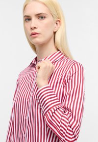 Camicia a righe rossa e bianca con colletto abbottonato, maniche lunghe e una texture di tessuto liscia. Il motivo presenta strisce verticali sottili.