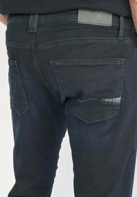 Jean en denim foncé avec une coupe standard, poches arrière avec des accents cousus et une étiquette en cuir à la taille. Texture lisse.