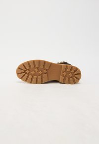 Semelle de botte marron avec des rainures profondes et des motifs en forme de croix sur un fond clair, montrant une adhérence texturée et le logo de la marque.
