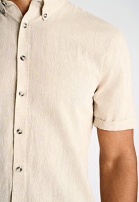 Beige kortharig knoopshirt met opgerolde manchetten, gedragen door een persoon met een gemiddelde huidtint, zichtbaar van schouder tot halverwege de romp.