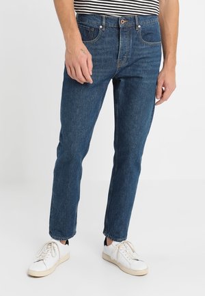 Homme portant un jean droit bleu foncé, des baskets blanches et une chemise rayée noir et blanc, debout sur un fond uni.