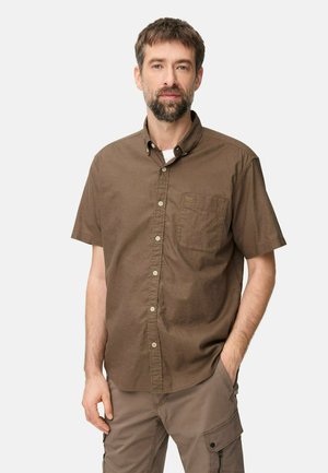 Mann mit Bart, der ein braunes kurzärmeliges Hemd mit Knöpfen und beigefarbene Cargohosen trägt, mit einer Hand in der Tasche, stehend vor einfarbigem Hintergrund.