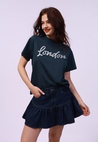 Camiseta de algodón verde oscuro con texto en plata "London", combinada con una mini falda de mezclilla oscura con dobladillo rizado y bolsillos.