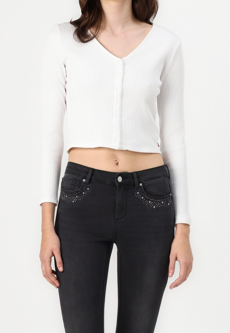 Weißes, geripptes, kurzes Langarmoberteil mit V-Ausschnitt und Knopfleiste, kombiniert mit schwarzen Jeans, die mit Nietenakzenten in den Taschen verziert sind.