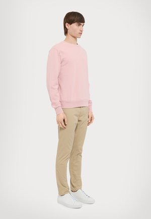 Sweatshirt rose clair avec des poignets et un ourlet côtelés, associé à un pantalon beige et des baskets blanches. Modèle se tenant de profil contre un fond uni.