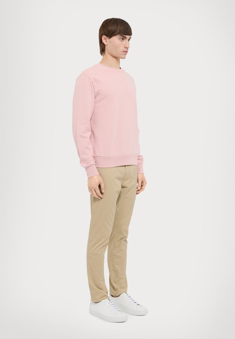 Lyserød sweatshirt med ribbede manchetter og kant, parret med beige bukser og hvide sneakers. Model stående på siden mod en ensfarvet baggrund.